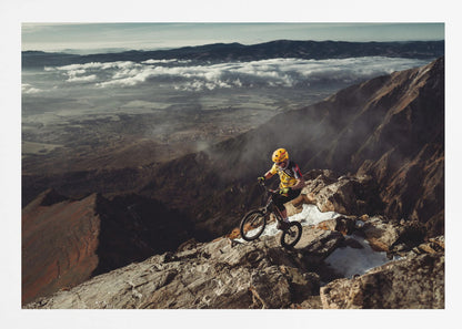 A mountain biker in a yellow helmet and jersey navigates a treacherous, rocky mountain ridge. Below, a vast valley is filled with a sea of clouds, with distant mountain ranges visible on the horizon. Print