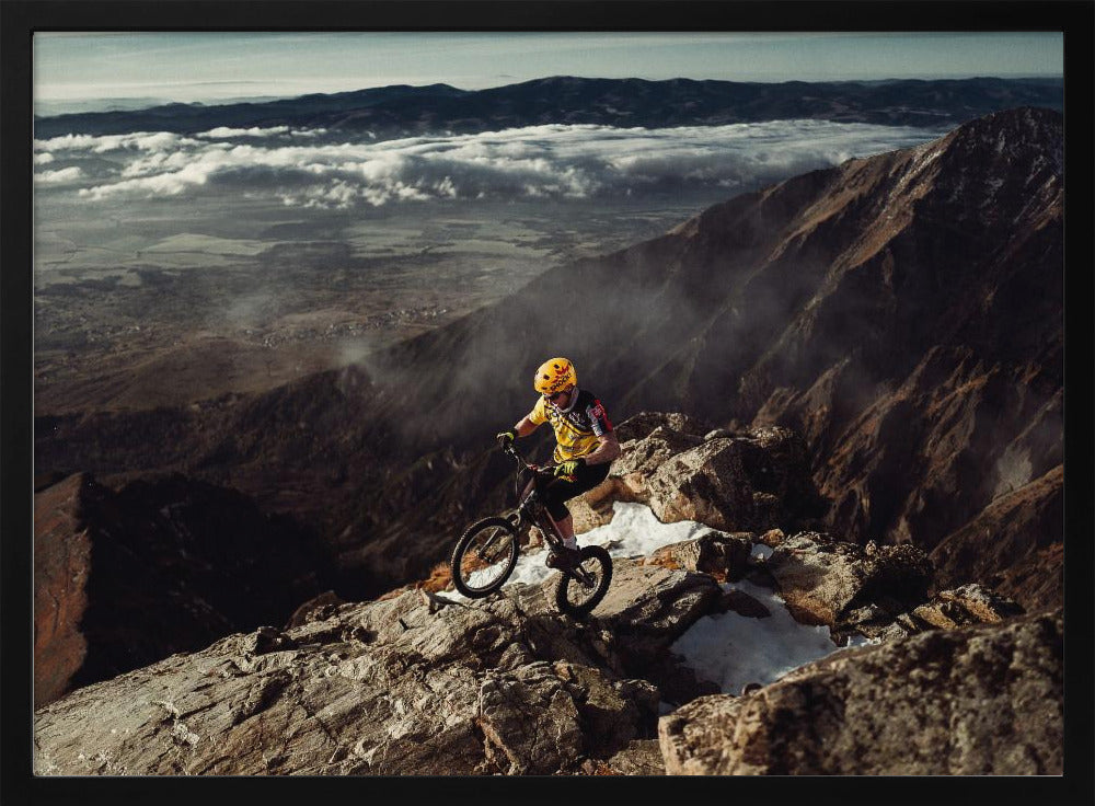A mountain biker in a yellow helmet and jersey navigates a treacherous, rocky mountain ridge. Below, a vast valley is filled with a sea of clouds, with distant mountain ranges visible on the horizon. Print