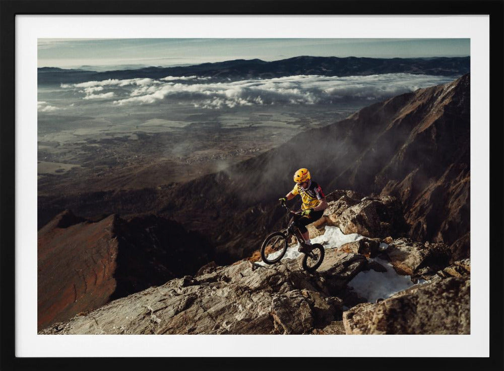 A mountain biker in a yellow helmet and jersey navigates a treacherous, rocky mountain ridge. Below, a vast valley is filled with a sea of clouds, with distant mountain ranges visible on the horizon. Print