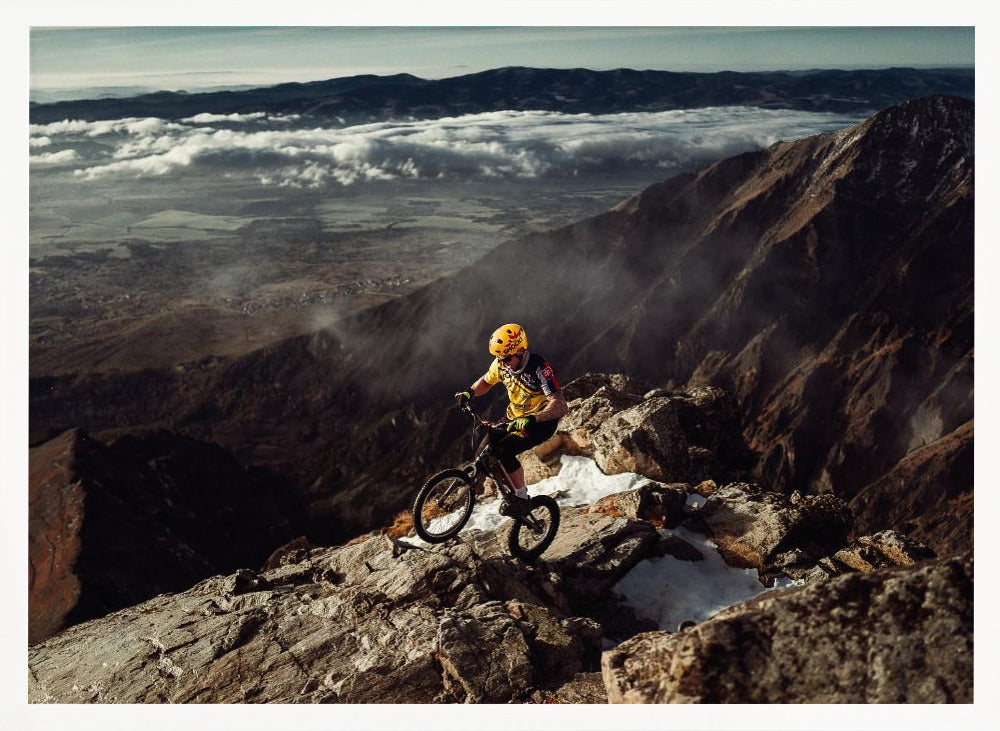 A mountain biker in a yellow helmet and jersey navigates a treacherous, rocky mountain ridge. Below, a vast valley is filled with a sea of clouds, with distant mountain ranges visible on the horizon. Print