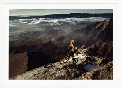 A mountain biker in a yellow helmet and jersey navigates a treacherous, rocky mountain ridge. Below, a vast valley is filled with a sea of clouds, with distant mountain ranges visible on the horizon. Print