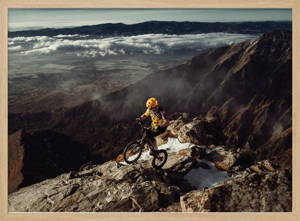 A mountain biker in a yellow helmet and jersey navigates a treacherous, rocky mountain ridge. Below, a vast valley is filled with a sea of clouds, with distant mountain ranges visible on the horizon. Print