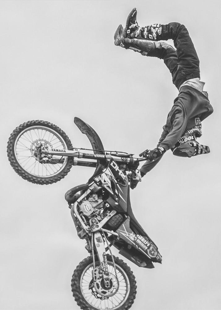 A dynamic black and white action shot of a freestyle motocross rider performing a handstand on the handlebars of their dirt bike mid-air. The bike is nearly vertical against a plain white sky, capturing a moment of extreme athleticism and balance. Poster