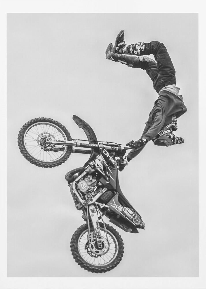 A dynamic black and white action shot of a freestyle motocross rider performing a handstand on the handlebars of their dirt bike mid-air. The bike is nearly vertical against a plain white sky, capturing a moment of extreme athleticism and balance. Poster