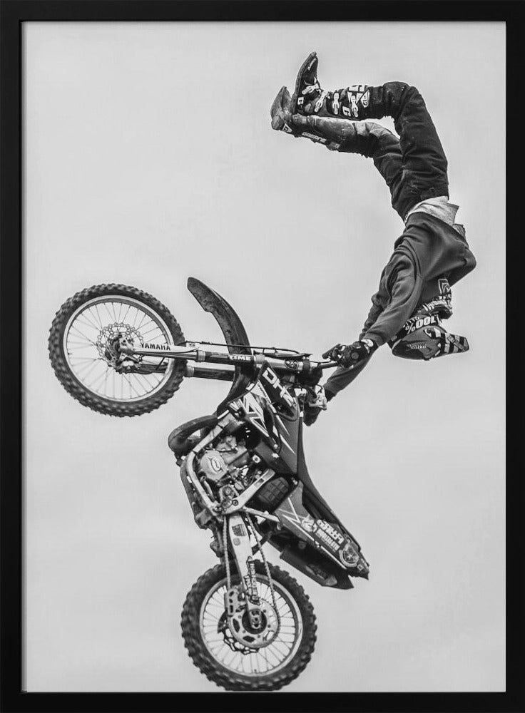 A dynamic black and white action shot of a freestyle motocross rider performing a handstand on the handlebars of their dirt bike mid-air. The bike is nearly vertical against a plain white sky, capturing a moment of extreme athleticism and balance. Poster