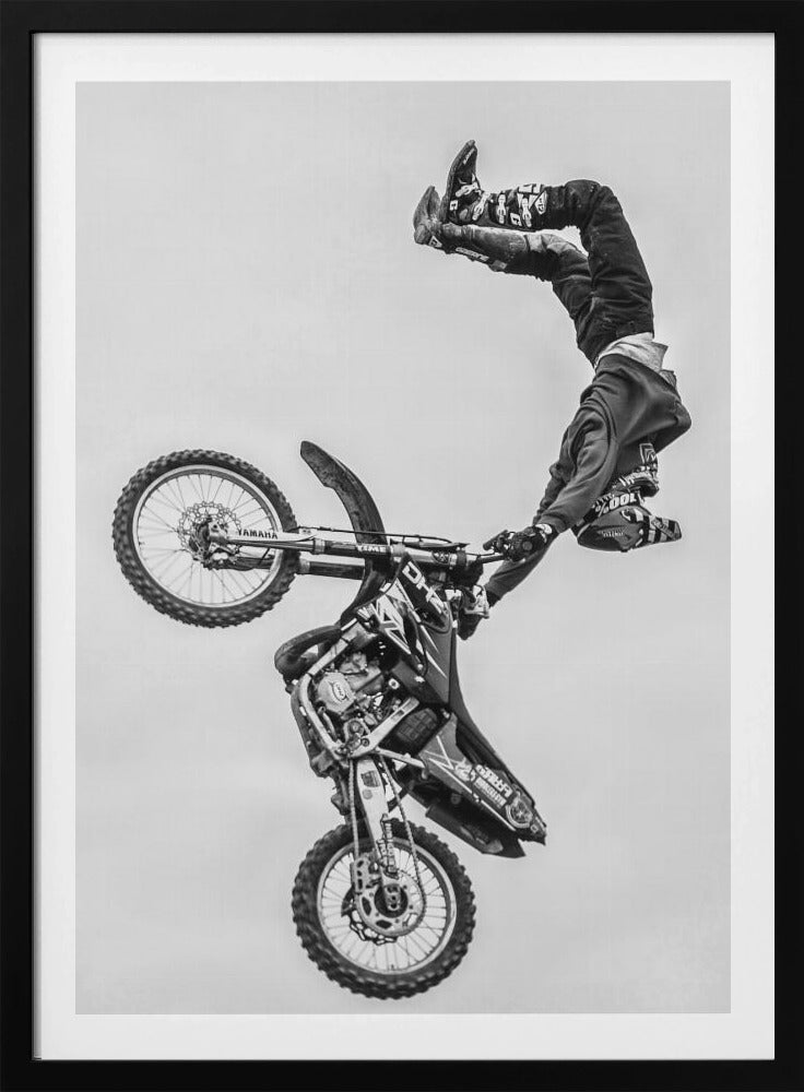 A dynamic black and white action shot of a freestyle motocross rider performing a handstand on the handlebars of their dirt bike mid-air. The bike is nearly vertical against a plain white sky, capturing a moment of extreme athleticism and balance. Poster