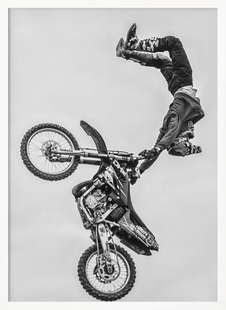 A dynamic black and white action shot of a freestyle motocross rider performing a handstand on the handlebars of their dirt bike mid-air. The bike is nearly vertical against a plain white sky, capturing a moment of extreme athleticism and balance. Poster