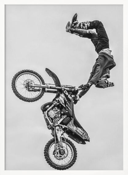 A dynamic black and white action shot of a freestyle motocross rider performing a handstand on the handlebars of their dirt bike mid-air. The bike is nearly vertical against a plain white sky, capturing a moment of extreme athleticism and balance. Poster