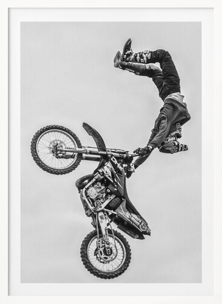 A dynamic black and white action shot of a freestyle motocross rider performing a handstand on the handlebars of their dirt bike mid-air. The bike is nearly vertical against a plain white sky, capturing a moment of extreme athleticism and balance. Poster