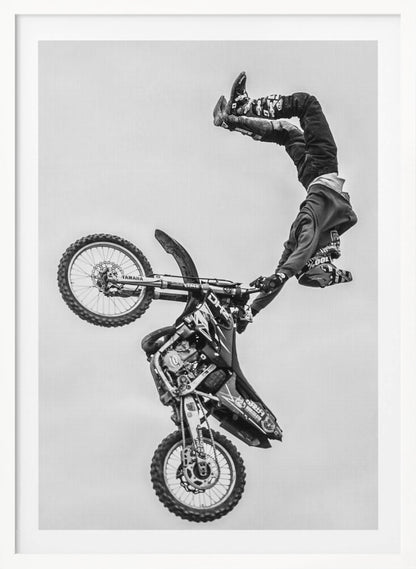 A dynamic black and white action shot of a freestyle motocross rider performing a handstand on the handlebars of their dirt bike mid-air. The bike is nearly vertical against a plain white sky, capturing a moment of extreme athleticism and balance. Poster