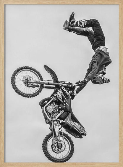 A dynamic black and white action shot of a freestyle motocross rider performing a handstand on the handlebars of their dirt bike mid-air. The bike is nearly vertical against a plain white sky, capturing a moment of extreme athleticism and balance. Poster