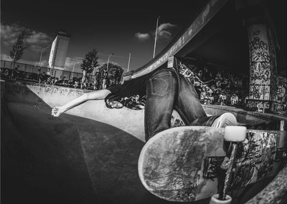 A dynamic, wide-angle black and white photograph of a skateboarder at a skatepark. The skater is bent backwards over the curved edge of a concrete bowl, with one hand touching the surface. Their skateboard is propped up in the foreground, and the background is filled with graffiti-covered walls, spectators, and a tall building against a dark, cloudy sky. Artwork