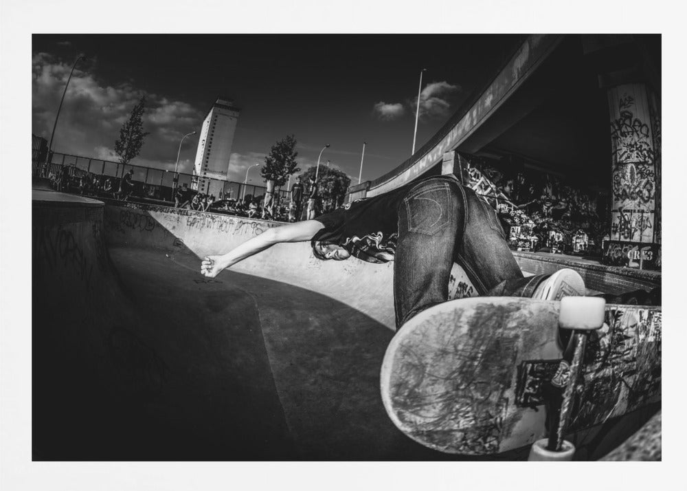 A dynamic, wide-angle black and white photograph of a skateboarder at a skatepark. The skater is bent backwards over the curved edge of a concrete bowl, with one hand touching the surface. Their skateboard is propped up in the foreground, and the background is filled with graffiti-covered walls, spectators, and a tall building against a dark, cloudy sky. Artwork
