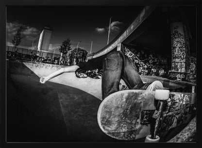A dynamic, wide-angle black and white photograph of a skateboarder at a skatepark. The skater is bent backwards over the curved edge of a concrete bowl, with one hand touching the surface. Their skateboard is propped up in the foreground, and the background is filled with graffiti-covered walls, spectators, and a tall building against a dark, cloudy sky. Artwork