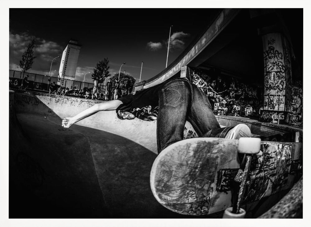 A dynamic, wide-angle black and white photograph of a skateboarder at a skatepark. The skater is bent backwards over the curved edge of a concrete bowl, with one hand touching the surface. Their skateboard is propped up in the foreground, and the background is filled with graffiti-covered walls, spectators, and a tall building against a dark, cloudy sky. Artwork