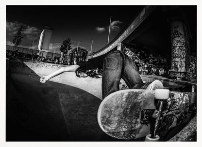 A dynamic, wide-angle black and white photograph of a skateboarder at a skatepark. The skater is bent backwards over the curved edge of a concrete bowl, with one hand touching the surface. Their skateboard is propped up in the foreground, and the background is filled with graffiti-covered walls, spectators, and a tall building against a dark, cloudy sky. Artwork