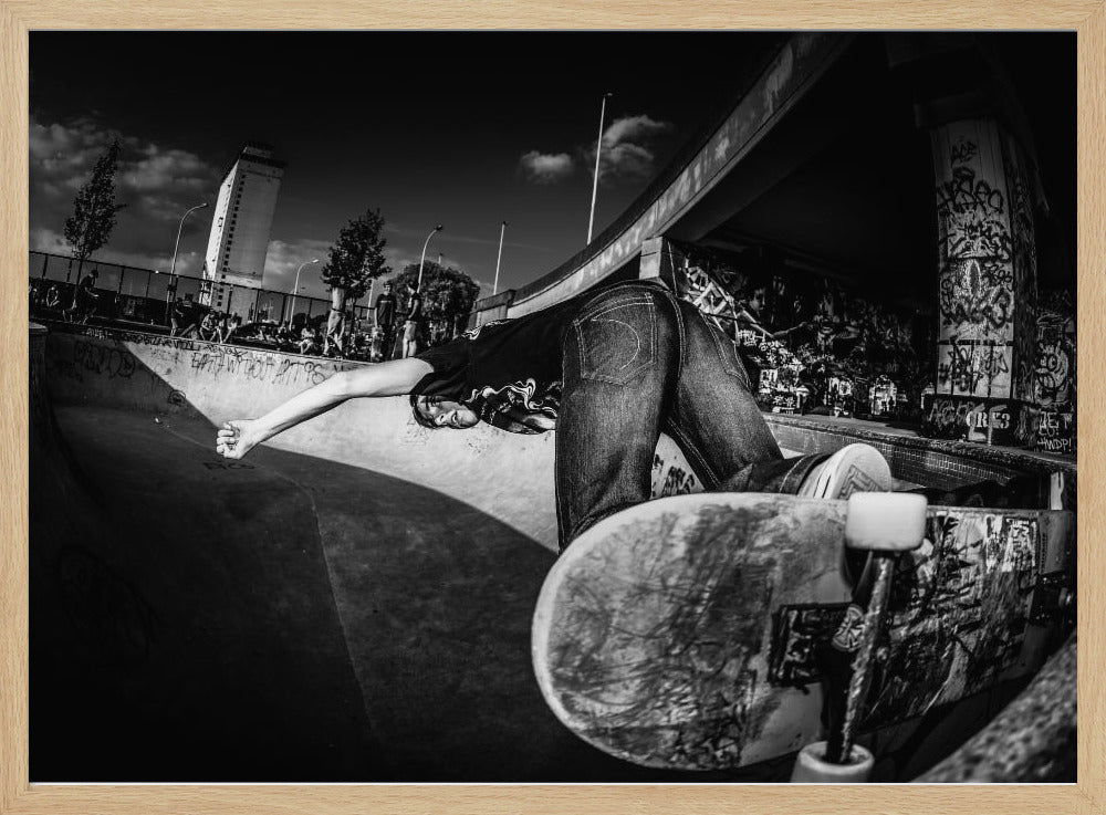 A dynamic, wide-angle black and white photograph of a skateboarder at a skatepark. The skater is bent backwards over the curved edge of a concrete bowl, with one hand touching the surface. Their skateboard is propped up in the foreground, and the background is filled with graffiti-covered walls, spectators, and a tall building against a dark, cloudy sky. Artwork