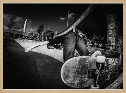 A dynamic, wide-angle black and white photograph of a skateboarder at a skatepark. The skater is bent backwards over the curved edge of a concrete bowl, with one hand touching the surface. Their skateboard is propped up in the foreground, and the background is filled with graffiti-covered walls, spectators, and a tall building against a dark, cloudy sky. Artwork