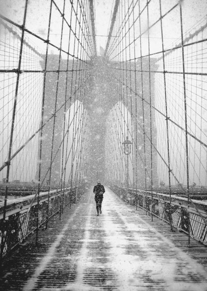 A vertical, black and white photograph of a solitary person walking along the Brooklyn Bridge during a heavy snowstorm. The iconic cables and towers of the bridge create a strong sense of perspective, drawing the eye towards the distant, hazy towers while snow falls all around. Artwork