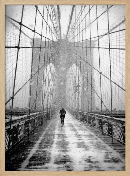 A vertical, black and white photograph of a solitary person walking along the Brooklyn Bridge during a heavy snowstorm. The iconic cables and towers of the bridge create a strong sense of perspective, drawing the eye towards the distant, hazy towers while snow falls all around. Artwork