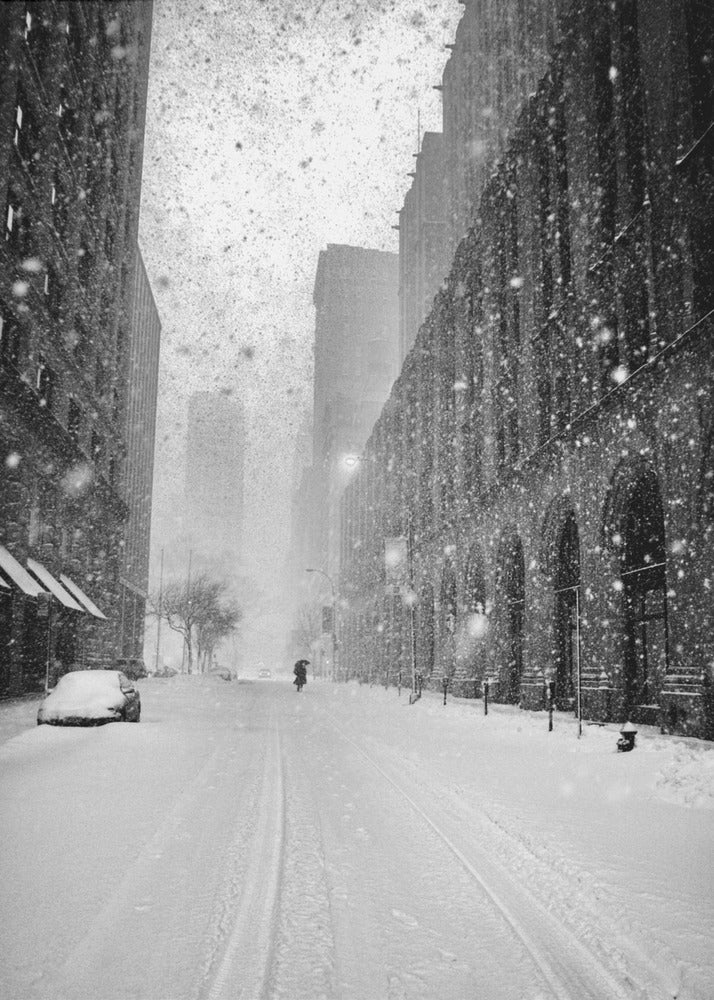 A grainy, black and white photograph captures a deserted city street during a heavy snowstorm. Tall buildings line the street, which is covered in a thick blanket of snow. A single car on the left is almost completely buried, and in the distance, a lone figure walks with an umbrella down the center of the empty road. Poster