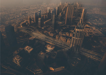 An aerial view of a sprawling modern city at dusk, with the warm glow of sunset casting long shadows over skyscrapers and illuminating a complex network of highways. The photograph has a warm, sepia-like tone and is presented within a silver-colored frame. Decor