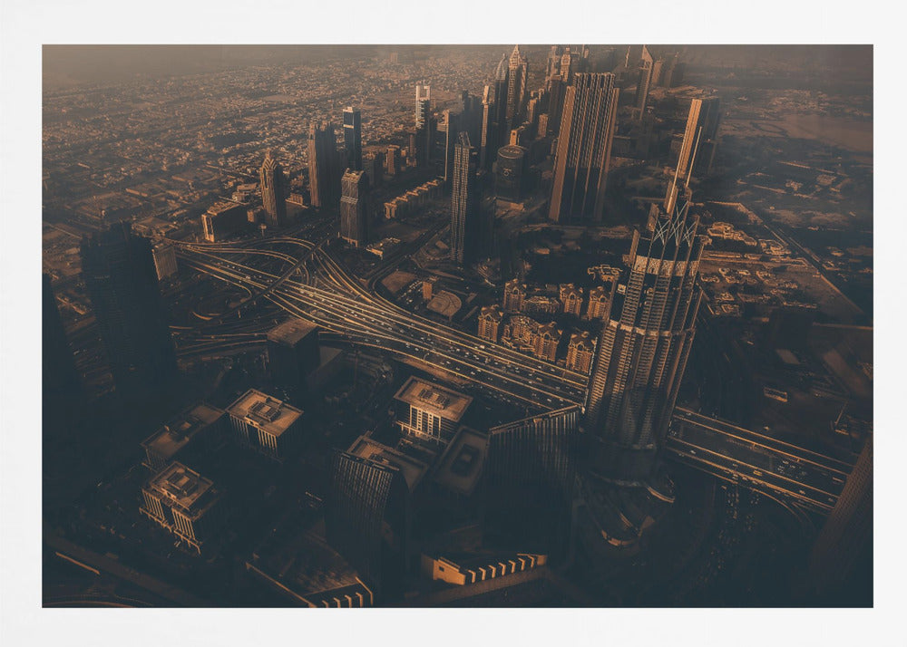 An aerial view of a sprawling modern city at dusk, with the warm glow of sunset casting long shadows over skyscrapers and illuminating a complex network of highways. The photograph has a warm, sepia-like tone and is presented within a silver-colored frame. Decor