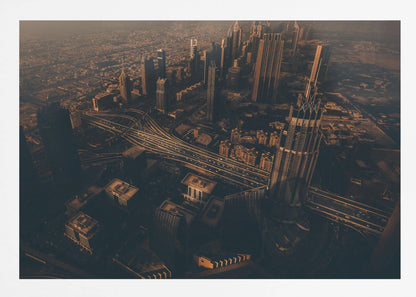 An aerial view of a sprawling modern city at dusk, with the warm glow of sunset casting long shadows over skyscrapers and illuminating a complex network of highways. The photograph has a warm, sepia-like tone and is presented within a silver-colored frame. Decor