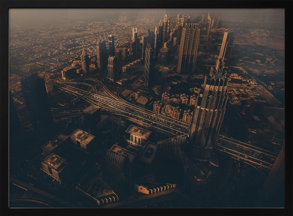 An aerial view of a sprawling modern city at dusk, with the warm glow of sunset casting long shadows over skyscrapers and illuminating a complex network of highways. The photograph has a warm, sepia-like tone and is presented within a silver-colored frame. Decor