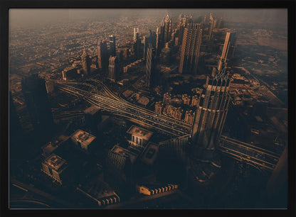 An aerial view of a sprawling modern city at dusk, with the warm glow of sunset casting long shadows over skyscrapers and illuminating a complex network of highways. The photograph has a warm, sepia-like tone and is presented within a silver-colored frame. Decor