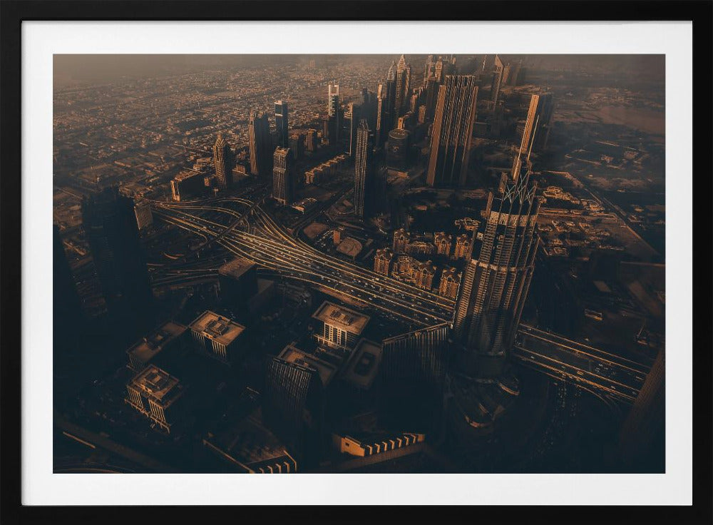 An aerial view of a sprawling modern city at dusk, with the warm glow of sunset casting long shadows over skyscrapers and illuminating a complex network of highways. The photograph has a warm, sepia-like tone and is presented within a silver-colored frame. Decor