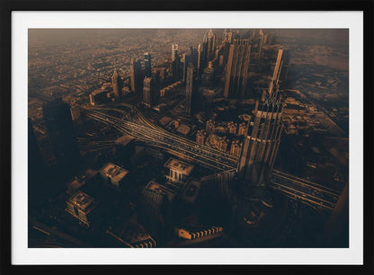 An aerial view of a sprawling modern city at dusk, with the warm glow of sunset casting long shadows over skyscrapers and illuminating a complex network of highways. The photograph has a warm, sepia-like tone and is presented within a silver-colored frame. Decor