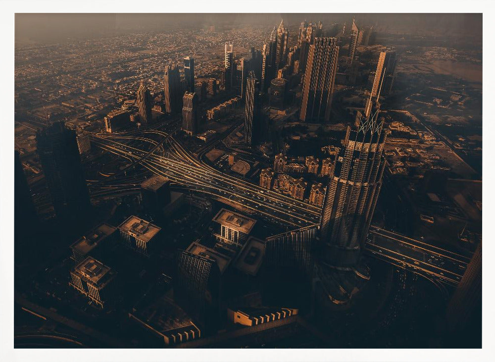 An aerial view of a sprawling modern city at dusk, with the warm glow of sunset casting long shadows over skyscrapers and illuminating a complex network of highways. The photograph has a warm, sepia-like tone and is presented within a silver-colored frame. Decor