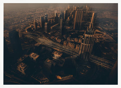 An aerial view of a sprawling modern city at dusk, with the warm glow of sunset casting long shadows over skyscrapers and illuminating a complex network of highways. The photograph has a warm, sepia-like tone and is presented within a silver-colored frame. Decor