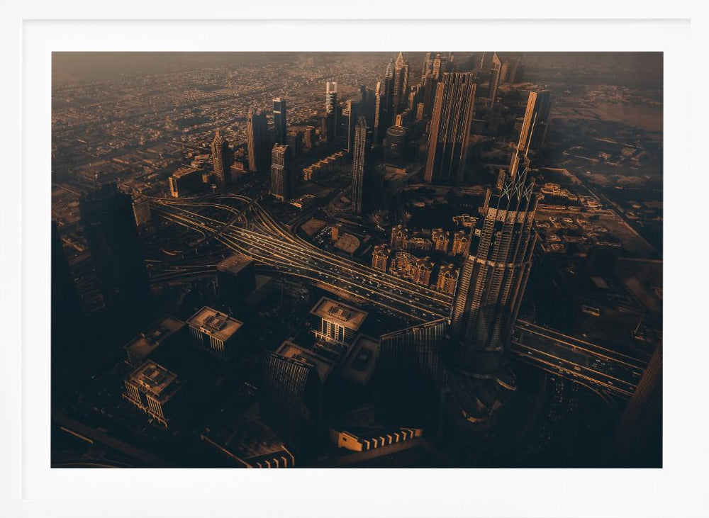 An aerial view of a sprawling modern city at dusk, with the warm glow of sunset casting long shadows over skyscrapers and illuminating a complex network of highways. The photograph has a warm, sepia-like tone and is presented within a silver-colored frame. Decor