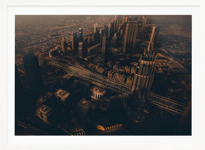 An aerial view of a sprawling modern city at dusk, with the warm glow of sunset casting long shadows over skyscrapers and illuminating a complex network of highways. The photograph has a warm, sepia-like tone and is presented within a silver-colored frame. Decor