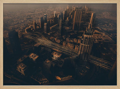 An aerial view of a sprawling modern city at dusk, with the warm glow of sunset casting long shadows over skyscrapers and illuminating a complex network of highways. The photograph has a warm, sepia-like tone and is presented within a silver-colored frame. Decor