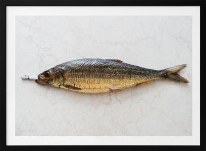 A surreal photograph of a whole smoked fish with a cigarette butt in its mouth, laying horizontally on a white marble surface, all within a silver frame. Wall Art