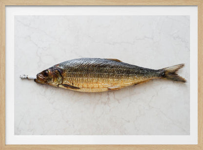 A surreal photograph of a whole smoked fish with a cigarette butt in its mouth, laying horizontally on a white marble surface, all within a silver frame. Wall Art