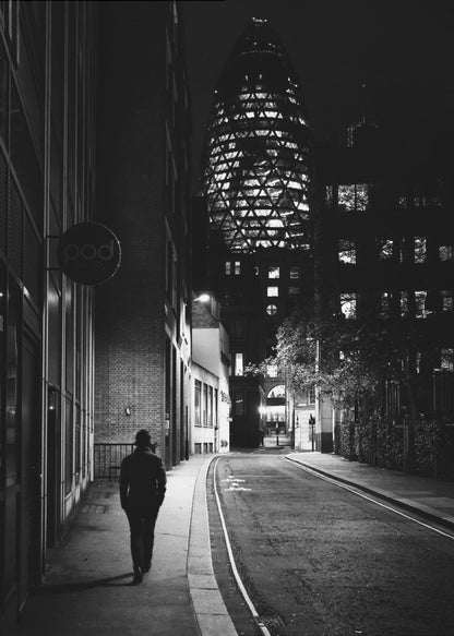 A moody black and white photograph of a person walking away down a narrow, deserted city street at night. In the background, the iconic Gherkin building in London is brightly lit against the dark sky. Wall Art
