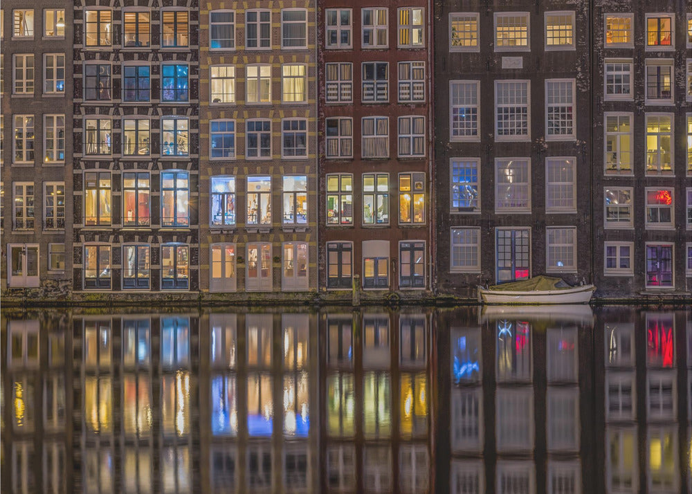 A framed nighttime photograph of a row of traditional Amsterdam canal houses with many illuminated windows, their colorful lights reflecting perfectly in the calm canal water below. A small white boat is moored at the water's edge. Decor