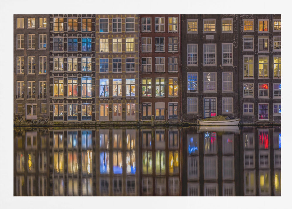 A framed nighttime photograph of a row of traditional Amsterdam canal houses with many illuminated windows, their colorful lights reflecting perfectly in the calm canal water below. A small white boat is moored at the water's edge. Decor