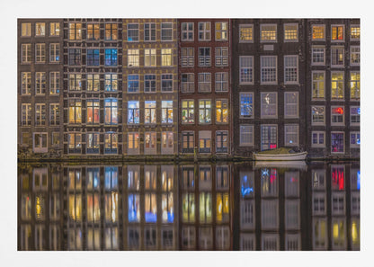 A framed nighttime photograph of a row of traditional Amsterdam canal houses with many illuminated windows, their colorful lights reflecting perfectly in the calm canal water below. A small white boat is moored at the water's edge. Decor