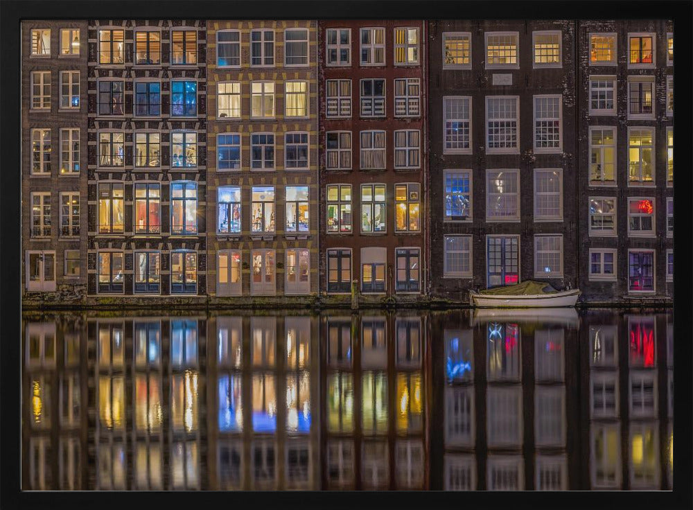 A framed nighttime photograph of a row of traditional Amsterdam canal houses with many illuminated windows, their colorful lights reflecting perfectly in the calm canal water below. A small white boat is moored at the water's edge. Decor
