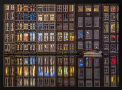 A framed nighttime photograph of a row of traditional Amsterdam canal houses with many illuminated windows, their colorful lights reflecting perfectly in the calm canal water below. A small white boat is moored at the water's edge. Decor