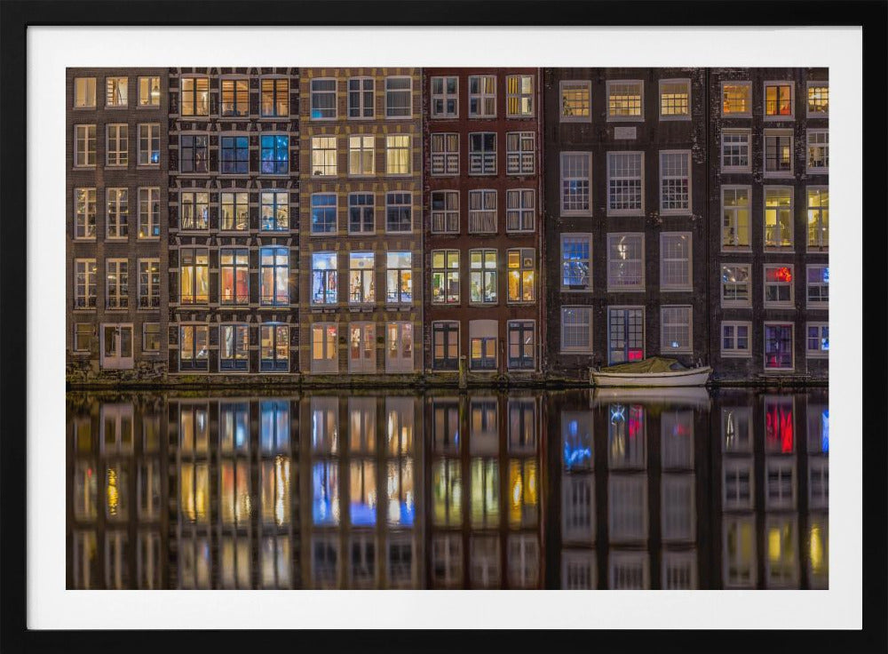 A framed nighttime photograph of a row of traditional Amsterdam canal houses with many illuminated windows, their colorful lights reflecting perfectly in the calm canal water below. A small white boat is moored at the water's edge. Decor