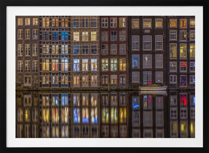 A framed nighttime photograph of a row of traditional Amsterdam canal houses with many illuminated windows, their colorful lights reflecting perfectly in the calm canal water below. A small white boat is moored at the water's edge. Decor