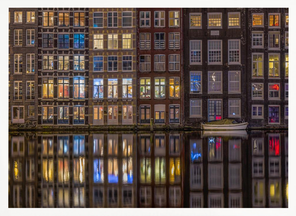 A framed nighttime photograph of a row of traditional Amsterdam canal houses with many illuminated windows, their colorful lights reflecting perfectly in the calm canal water below. A small white boat is moored at the water's edge. Decor