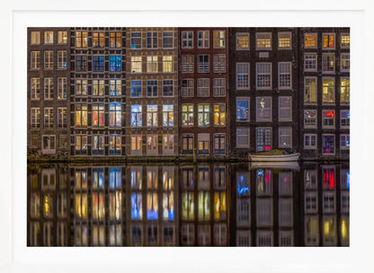 A framed nighttime photograph of a row of traditional Amsterdam canal houses with many illuminated windows, their colorful lights reflecting perfectly in the calm canal water below. A small white boat is moored at the water's edge. Decor