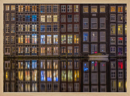 A framed nighttime photograph of a row of traditional Amsterdam canal houses with many illuminated windows, their colorful lights reflecting perfectly in the calm canal water below. A small white boat is moored at the water's edge. Decor